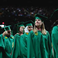 A group of graduates wearing caps and gowns look hopefully up into the stands at EagleBank Arena on convocation day
