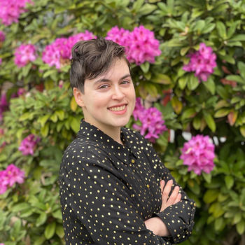 CJ Mandell A photo of CJ with arms crossed and angled to the right in front of a slightly blurred background of a green leafy plant with bright pink bunches of flowers. They are wearing a black collared long-sleeve shirt with a pattern of gold dots and buttons and smiling while making eye contact with the camera. 